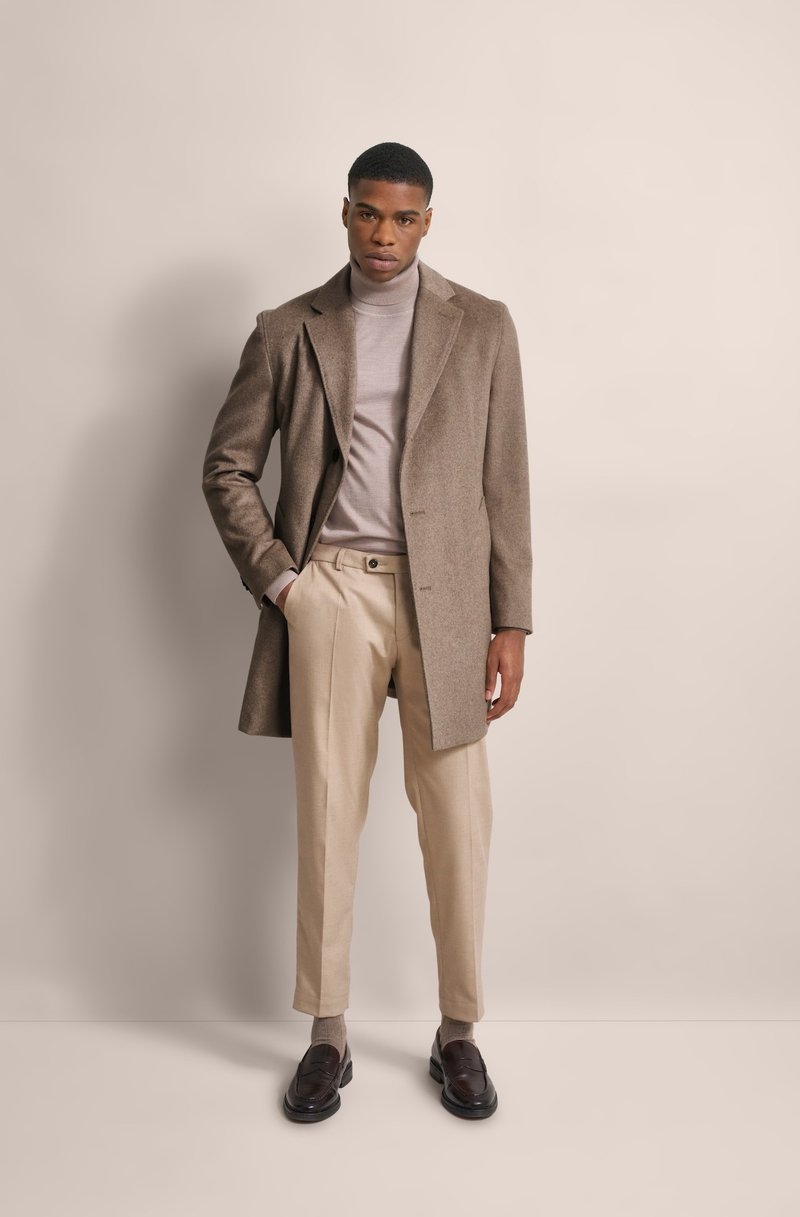 Cappotto lungo in misto lana marrone sopra un leggero dolcevita, abbinato a pantaloni beige; uomo in piedi con le mani in tasca, indossa mocassini scuri.