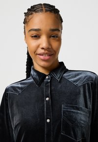 Camicia in velluto nero con colletto a punta, bottoni in perla e taschino sul petto con cuciture a contrasto e dettagli sulle spalle.