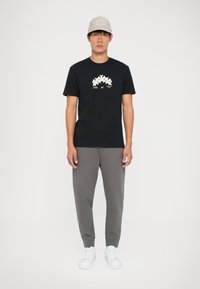 Schwarzes Baumwoll-T-Shirt mit einem grafischen Design in Weiß. Kombiniert mit grauen Jogginghosen und weißen Sneakern. Modell trägt eine hellgraue Mütze.