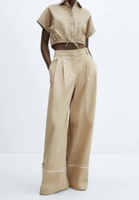 Una donna indossa una camicia a pulsanti cropped a maniche corte beige e pantaloni a gamba larga a vita alta con orli cuff e bordi bianchi, in piedi con le mani in tasca.