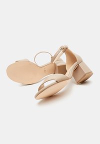 Sandalias de tacón beige con diseño de correas cruzadas, punta descubierta, correa ajustable en el tobillo y tacón cuadrado. Material de cuero liso con acabado mate.
