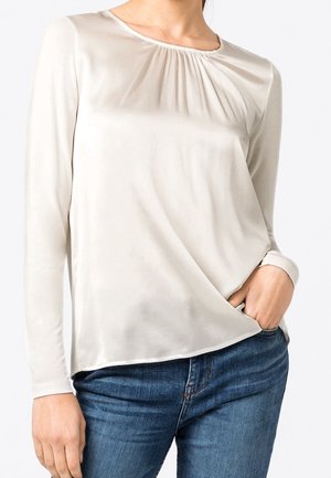 Vrouw die een crèmekleurige blouse met lange mouwen draagt, met subtiele plooien bij de kraag, gecombineerd met een blauwe spijkerbroek, hand in de zak.