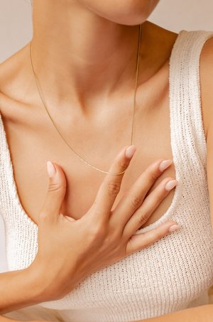 Collana sottile d'oro posata su una maglia bianca testurizzata. Le unghie sono laccate di un colore chiaro. Il tono della pelle appare uniforme e liscio.