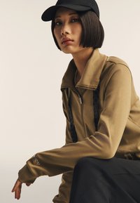 Chaqueta de color caqui con cremalleras y detalles en negro; mangas largas con agujeros para los pulgares. El modelo lleva una gorra negra y tiene un peinado con corte recto.