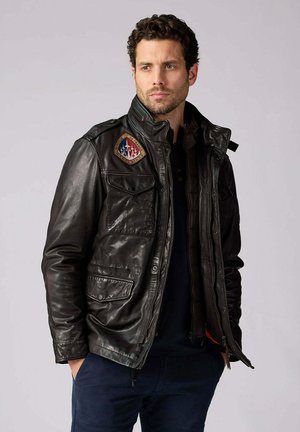 Homme aux cheveux courts et bouclés portant une veste en cuir noire boutonnée avec plusieurs poches et un écusson coloré sur la poitrine, les mains dans les poches.