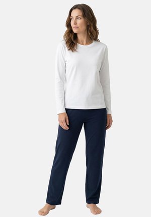 Vrouw staat blootsvoets, draagt een wit shirt met lange mouwen en losse marineblauwe broek, kijkt naar de zijkant tegen een effen achtergrond.