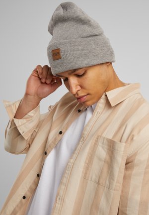 Gray knit beanie with a square leather patch. Striped beige long-sleeve shirt with black buttons and a breast pocket. White T-shirt underneath.