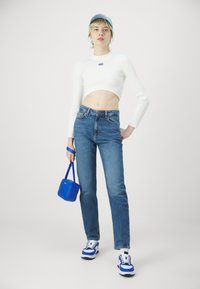 Geribbelde witte cropped top, blauwe high-waisted jeans, blauwe handtas en blauw-witte sneakers, met een lichtblauwe pet en een rechte pasvorm.