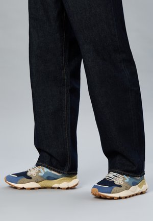 Gambe con jeans blu scuro e sneakers multicolori con accenti beige, blu e navy, in piedi su un pavimento grigio chiaro.