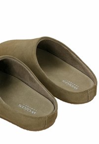 Olijfgroene slippers met een zachte suède buitenkant, gevoerd voetbed, ronde vorm en minimale stikseldetails. Merkenlogo zichtbaar aan de binnenkant.