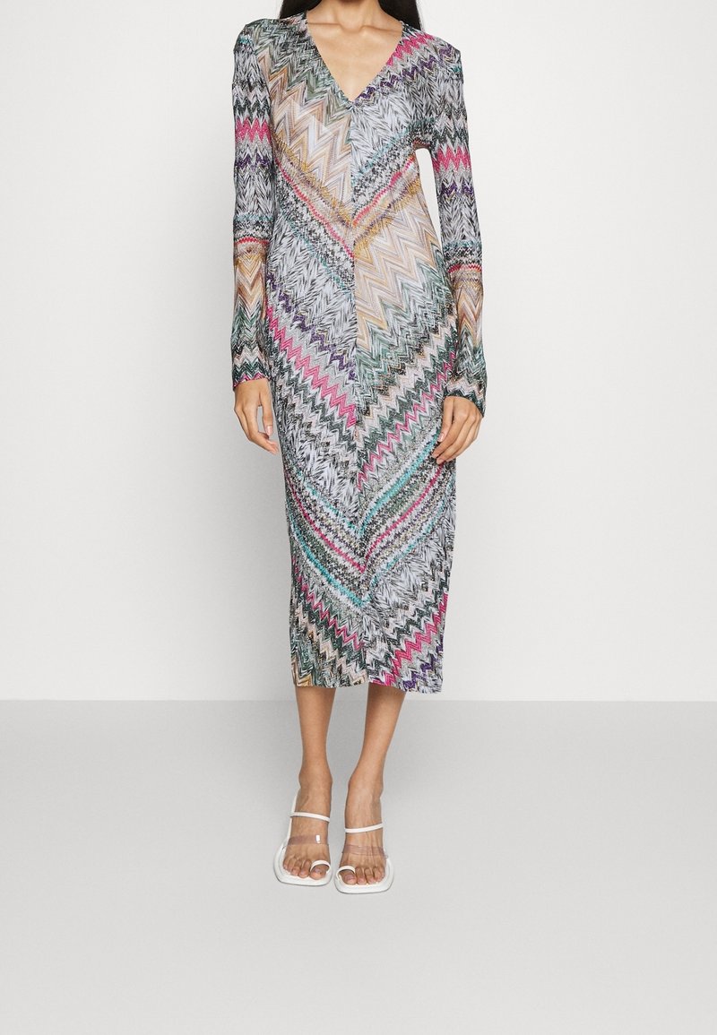 Femme portant une robe midi à manches longues et col en V avec des motifs zigzag colorés, associée à des talons blancs à bouts ouverts sur un fond uni.