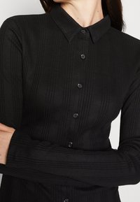 Chemise noire côtelée avec col pointu, manches longues et fermeture à boutons. Présente une texture verticale subtile et des boutons sur le devant.