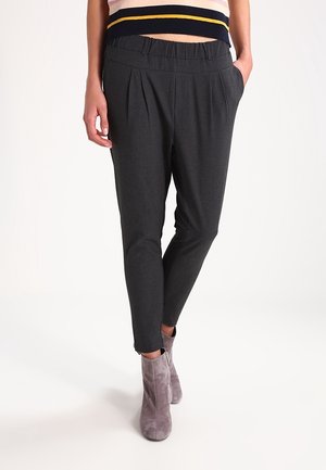 Femme portant un pantalon fuselé gris foncé avec des plis et des bottines en daim gris, main dans la poche, top sans manches rayé visible coupé au-dessus.