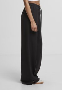 Fekete, bő szárú nadrág, húzózsinórral a derékrészen és oldalsó szellőzőkkel. Sima anyagból készült, laza látványú. Az alján látható egy pár sportcipő.