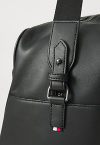Black synthetic leather bag with a textured finish, featuring a metal buckle, adjustable strap, and a small tricolor logo at the bottom.