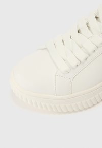 Fehér sportcipő sima bőr felsőrésszel, kerek orr-résszel, lapos fehér fűzőkkel és apró bordázattal ellátott texturált gumitalppal.