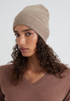 Cappello a maglia marrone con texture a coste e punta rastremata, realizzato in materiale morbido. Indossato con un maglione a scollo a V dello stesso tono.