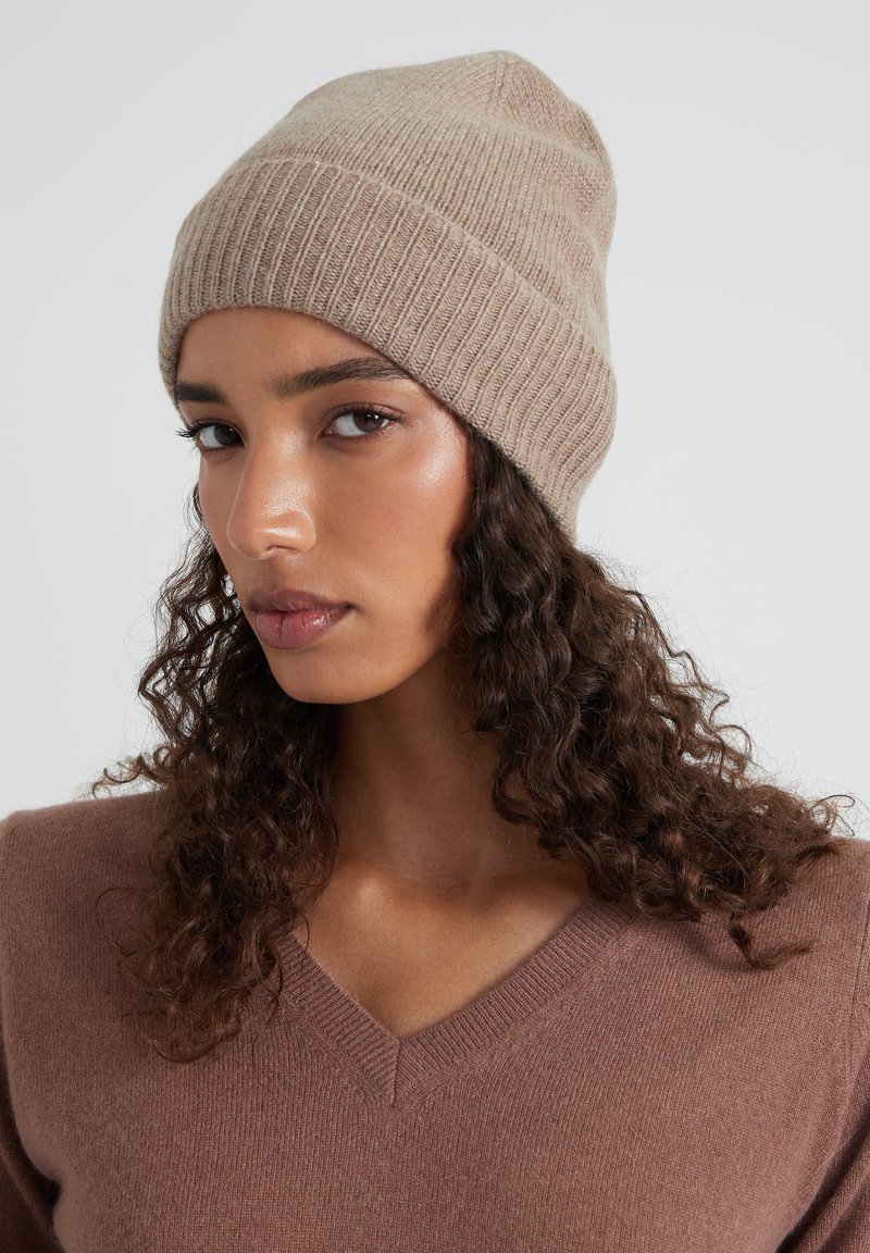 Bonnet en laine marron avec une texture côtelée et un sommet effilé, fabriqué en matériau doux. Porté avec un pull à col en V de couleur assortie.