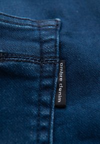 Mørkeblå denimstof med synlig tekstur. Indeholder syningsdetaljer og et lille sort mærke med hvid tekst, der lyder "ombre denim."