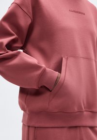 Άτομο που φοράει φούτερ χρώματος σκόπιου ροζ με ριμπ μανσέτες και τελείωμα, χέρι στην μπροστινή τσέπη και διακριτικό όνομα εμπορικής μάρκας στο στήθος.