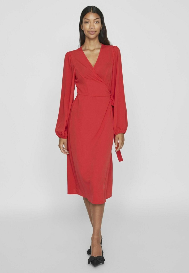 Červené šaty s dlhými nafúknutými rukávmi a viazaním v páse. Hladká látka, dĺžka po kolená, nosené s čiernymi topánkami na mašľu a vysokom opätku.