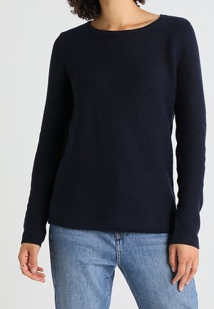 Kvinde iført en tætsiddende, langærmet navyblå ribstrikket sweater kombineret med lyseblå denimjeans, stående foran en ensfarvet baggrund.