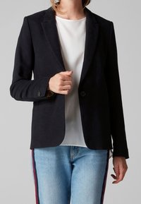 Blazer noir ajusté avec un revers cranté, deux boutons et poches avant, en tissu texturé ; porté sur une chemise blanche et un jean.