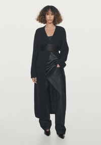 Lange zwarte cardigan gemaakt van zacht, donzig materiaal, met een diepe V-hals, brede mouwen en een ceintuur om de taille, gedragen over een satijnen top en denim broek.