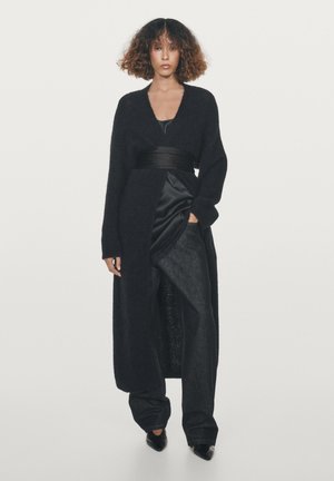 Lange zwarte cardigan gemaakt van zacht, donzig materiaal, met een diepe V-hals, brede mouwen en een ceintuur om de taille, gedragen over een satijnen top en denim broek.