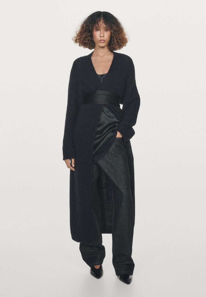 Lange zwarte cardigan gemaakt van zacht, donzig materiaal, met een diepe V-hals, brede mouwen en een ceintuur om de taille, gedragen over een satijnen top en denim broek.