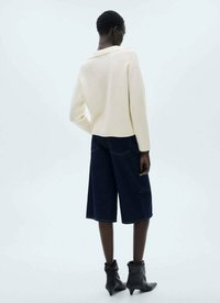 Crème ribbel gebreide trui met een uitgesneden kraag, gecombineerd met donkere denim culottes. Model draagt zwarte enkellaarsjes. Neutrale achtergrond.
