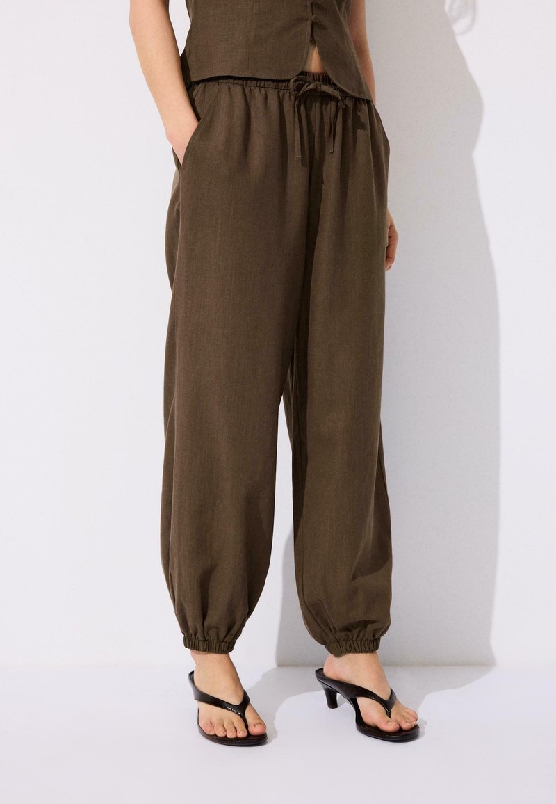 Femme portant un pantalon marron large avec cordon de serrage et poignets élastiques, ainsi que des sandales à talons noires, debout sur fond blanc.