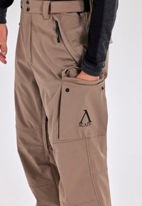 Beige cargobroek met een gestructureerde afwerking, voorzakken en een externe zak met een logo. Inclusief zwarte ritsdetails en knopen.