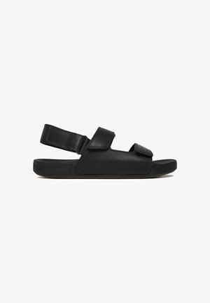 Sort åben sandal med justerbare remme over tæerne og anklen, flad sål og minimalistisk design.