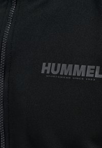 Svart sportjacka med dragkedja, med en mjuk textur och Hummel-logotyp i grått. Utformad för komfort och funktionalitet.