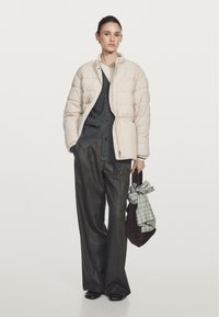 Giubbotto puffer beige chiaro con design trapuntato, cardigan grigio sottostante, pantaloni larghi scuri e una borsa marrone con una sciarpa a quadri.