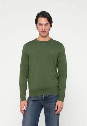 Pullover verde caratterizzato da un collo a giro, polsini e orlo a costine, con un piccolo logo sul petto. Indossato con jeans blu.