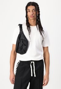 Black nylon sling bag featuring a smooth texture, angular shape, and zippered pocket. Worn over a white shirt and black pants with white accents.