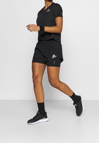 Černý sportovní outfit s přiléhavým topem a šortkami, vyrobený z lehkého materiálu, s jemným logem značky v bílé barvě.