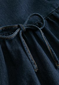 Tumša džinsa audums ar struktūras virsmu, kas satur sasietu loku un kontrastējošu dūrienu gar jostasvietu.