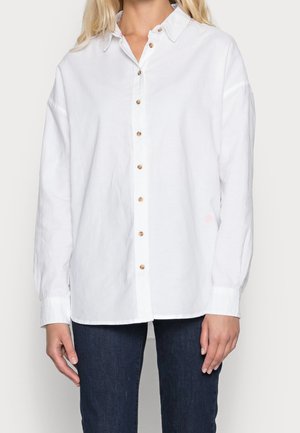 Femme portant une chemise blanche ample à boutons en bois et un jean bleu foncé, debout devant un fond uni.