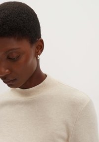 Beige gestreifter Pullover mit hohem Kragen, der eine subtile Textur aufweist. Einfaches Design, kombiniert mit kleinen silbernen Creolen.