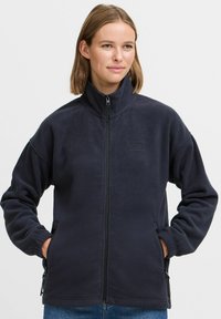 Marinefarget fleecejakke med høy krage, hel glidelås foran og glidelåslommer på siden. Myk tekstur, avslappet design og brodert logo.