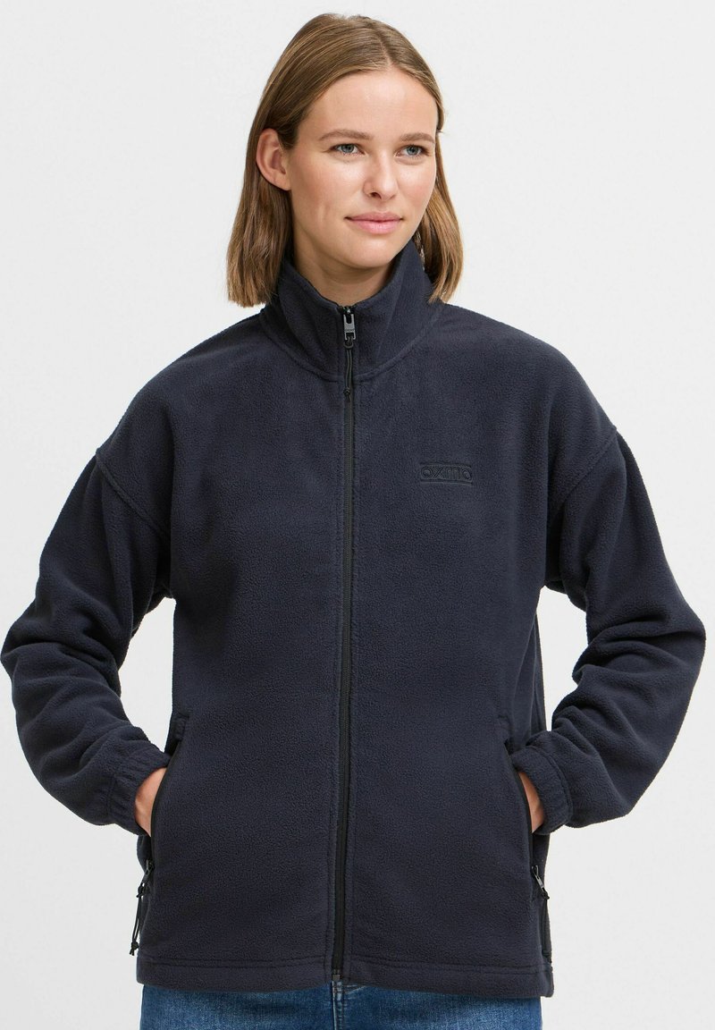 Marinefarget fleecejakke med høy krage, hel glidelås foran og glidelåslommer på siden. Myk tekstur, avslappet design og brodert logo.