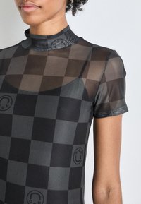 Zwart en grijs geruite mesh top met een hoge hals, korte mouwen en subtiele logo-details. Heeft een semi-doorzichtige textuur.