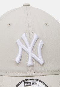 Beige baseball cap made of fabric with a raised white embroidered New York Yankees logo on the front and a black New Era tag attached.