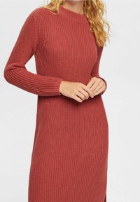 Robe pull en maille de couleur rouille, avec un col rond, des manches longues et une texture côtelée. Longueur sous le genou avec des fentes latérales.