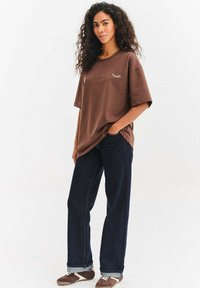 Brązowy oversize t-shirt z białą grafiką rzęsy, w połączeniu z ciemnoniebieskimi jeansami o prostych nogawkach oraz brązowymi sneakersami z białymi detalami.