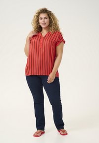Blusa de manga corta a rayas rojas con una fila de botones, combinada con vaqueros azul oscuro y chanclas rojas, sobre un fondo neutro.