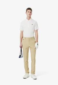 Hvid polo shirt med tre knapper og grønt logo, beige bukser, hvide sko, og en mesh taske med golfbolde i den ene hånd, iført en hvid handske.